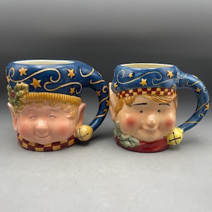 May include: Two ceramic mugs shaped like elf faces, ideal for festive occasions. Each mug features a smiling elf face with rosy cheeks, pointed ears, and a blue hat with gold stars. The handles are curved and have a small gold bell.