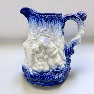May include: A blue and white ceramic pitcher with a detailed relief design. The pitcher features a tree-like handle and a base with a blue and white color scheme. The body of the pitcher has a relief of figures. The pitcher is a decorative item.