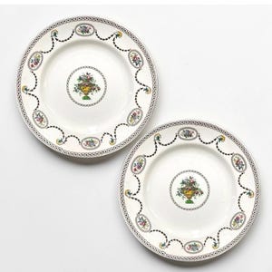 May include: Two white ceramic plates featuring a central illustration of a yellow urn filled with colorful flowers. The rims are decorated with a repeating pattern of small floral medallions connected by black swags and a dotted border design.