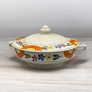 Può includere: Un piatto da portata in ceramica bianca con coperchio, con un motivo floreale in tonalità arancio, giallo, blu, verde e rosso. Il piatto ha due manici e un bordo scanalato.
