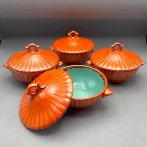 Könnte beinhalten: Ein Set aus vier runden Keramikschalen mit Deckeln, die eine leuchtend orangefarbene Außenseite mit einer gerippten, muschelartigen Textur und eine kontrastierende helltürkise Innenseite aufweisen. Jede Schale hat zwei kleine Seitengriffe.