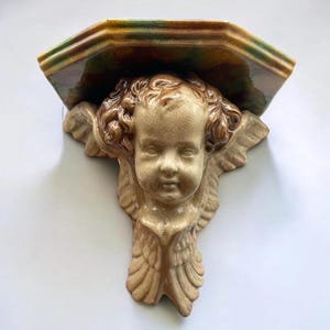 May include: A ceramic wall shelf featuring a sculpted cherub face with curly hair and wings. The shelf has a smooth, glossy finish with a beige and brown color palette, accented by green tones on the top surface and a visible crackle glaze texture.