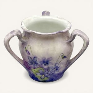 May include: A hand-painted, antique sugar bowl with a lid and two handles. The ceramic bowl is painted in shades of lavender and white, with a cluster of purple violets on the front. The interior is white.
