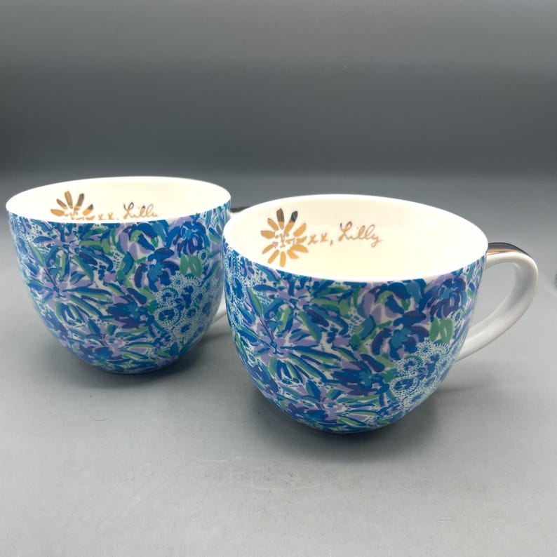 Puede incluir: Dos tazas de caf&eacute; de cer&aacute;mica con un estampado floral azul, verde y morado. Las tazas tienen un interior blanco con texto dorado que dice "xx, Lilly". Cada taza tiene un asa blanca.
