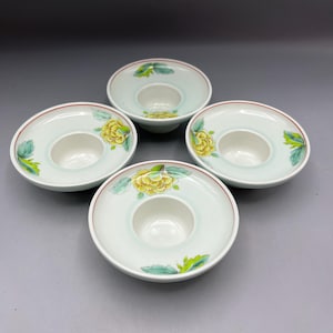 May include: Four white ceramic candle holders with a central well and a wide, flat rim. Each holder features a yellow floral design with green leaves and a red rim. The holders are arranged in a square formation against a gray background.