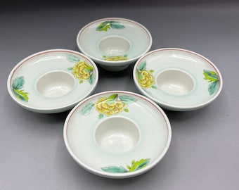 May include: Four white ceramic candle holders with a central well and a wide, flat rim. Each holder features a yellow floral design with green leaves and a red rim. The holders are arranged in a square formation against a gray background.