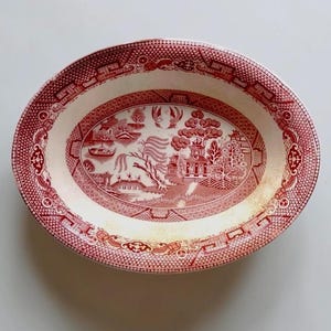 May include: Oval serving platter with a red and white Willow pattern. The platter features a detailed landscape scene in the center and a decorative border. The dish is made of ceramic and is suitable for serving food.
