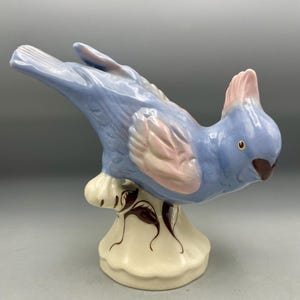 Puede incluir: Una figura de pájaro de cerámica con un cuerpo azul brillante y acentos de color rosa suave en las alas y la cresta. El pájaro está posado sobre una base de color crema decorada con patrones de hojas marrón oscuro y acabado craquelado.