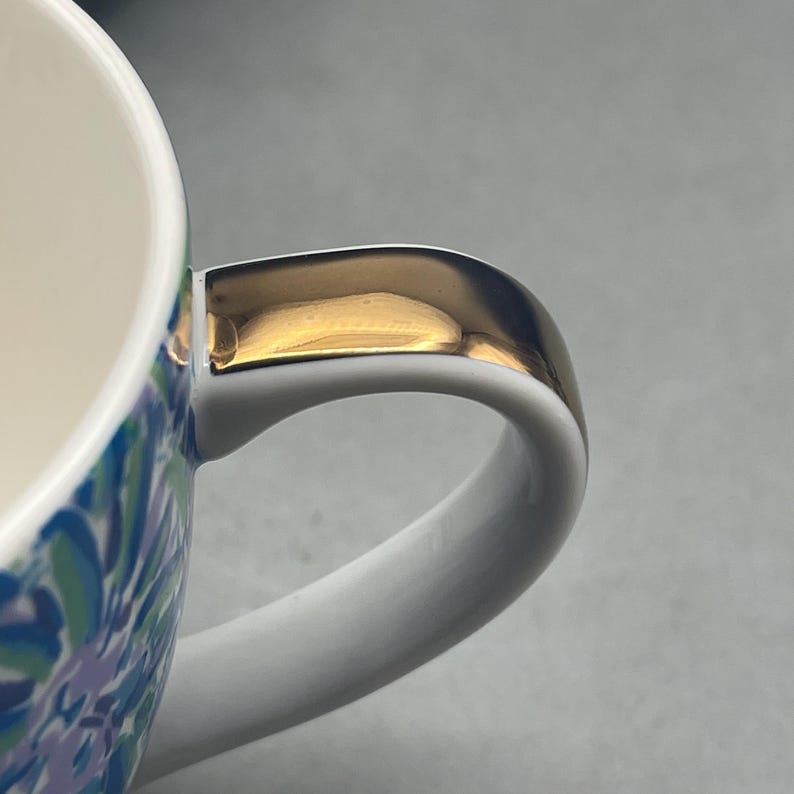 Puede incluir: Primer plano de una taza de cer&aacute;mica blanca con un asa dorada. El exterior de la taza presenta un estampado floral azul, verde y blanco. El asa es curva y tiene un acento dorado en la parte superior.