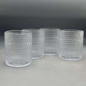 Pode incluir: Um conjunto de quatro copos de vidro transparente, cada um com uma superfície texturizada com faixas horizontais de pontos circulares em relevo de vários tamanhos. Os copos cilíndricos têm uma forma uniforme e acabamento límpido.