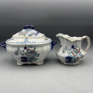 Puede incluir: Una fuente de cerámica blanca con tapa y una cremaera a juego. Ambas piezas están decoradas con motivos florales en tonos azules, rojos y verdes, con ribetes dorados. La fuente tiene asas azules.