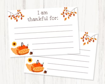 May include: Two white A6-sized Thanksgiving-themed printable cards. The cards have the text "I am thankful for:" with lined spaces for writing. They feature fall-themed illustrations of pumpkins, sunflowers, and cornucopias with autumn leaves.