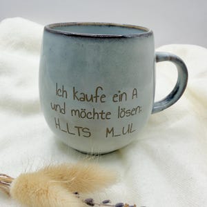 Könnte beinhalten: Eine hellblaue Keramik-Tasse mit dunklem Rand und Henkel. Die Tasse trägt den Text "Ich kaufe ein A und möchte lösen: H_LTS M_UL" in brauner Schrift. Die Tasse steht auf einer weißen Oberfläche.