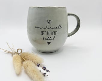 May include: A light gray ceramic mug with a rounded shape and a handle. The front features the black printed text "WIE wundervoll BIST DU DENN bitte?" followed by a small black heart symbol. The mug has a smooth, glazed finish.