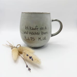 Könnte beinhalten: Ein hellgrauer Keramikbecher in abgerundeter Form mit Henkel, bedruckt mit dem schwarzen Text Ich kaufe ein A und möchte lösen: H_LTS M_UL. Vor dem Becher liegt ein kleines Bündel aus getrocknetem beigem Gras und violetten Lavendelzweigen.