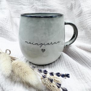 Könnte beinhalten: Ein hellgrauer Keramikbecher mit dunklem Rand und Henkel, der auf der Vorderseite den kursiven Schriftzug "moingiorno" und ein kleines Herzsymbol in Schwarz aufweist. Der Becher hat eine abgerundete, bauchige Form und eine glatte Oberfläche.