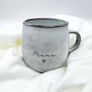 Könnte beinhalten: Ein weißer Keramikbecher mit abgerundeter Form und einem dunklen, strukturierten Rand. Das Wort Mama ist in schwarzer Schreibschrift auf der Vorderseite aufgedruckt, über einem kleinen, einfachen schwarzen Herzsymbol. Der Becher hat einen Henkel.