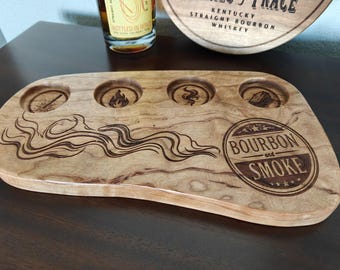 May include: A wooden serving tray with four circular cutouts and engraved designs. The tray features the text "Bourbon and Smoke" in a circular emblem. The wood has a natural grain and a light brown color.