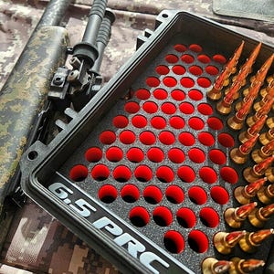 May include: A black case with red circular cutouts holds several gold-tipped bullets. The case is labeled "6.5 PRC" in white lettering. A camouflage rifle is partially visible in the background.