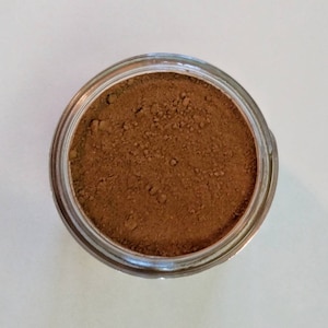 May include: Overhead view of a clear glass jar filled with a fine, brown powder. The powder has a slightly textured appearance and fills the jar completely. The jar's rim is visible, and the background is a plain white surface.