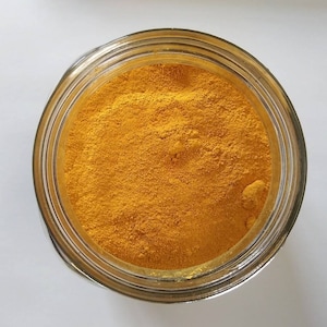 May include: Overhead view of a clear glass jar filled with bright yellow turmeric powder. The jar is open, revealing the finely ground spice. The background is a plain white surface, providing a clean contrast to the vibrant color of the turmeric.