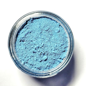 May include: A clear, circular glass jar filled with a fine, powdery substance in a vibrant sky blue color. The powder has a matte, textured appearance and fills the container to the brim, showing a consistent pigment throughout the surface.