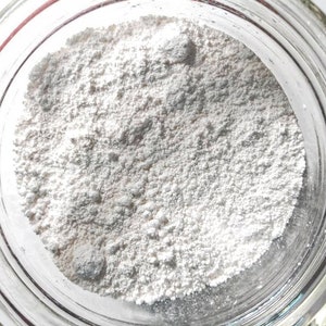 May include: A clear glass jar filled with a fine, off-white powder. The powder has a slightly textured appearance and fills the jar to the top. The jar's rim is visible, and the light reflects off the glass.