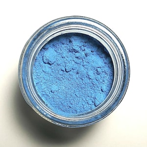 May include: A clear glass jar filled with a vibrant, finely milled sky blue powder. The container is viewed from directly above, showcasing the textured, matte surface of the pigment contained within the circular opening of the glass vessel.