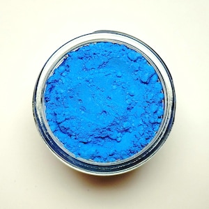 May include: Overhead shot of a small, clear glass jar filled with vibrant blue powder. The powder has a textured, granular appearance, filling the jar to the brim. The jar sits on a plain, off-white surface.