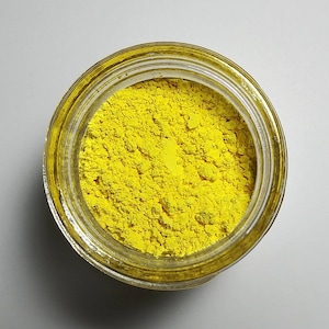 May include: A clear glass jar filled with bright yellow powder. The jar is open, revealing the finely ground substance inside. The powder has a textured appearance, and the jar sits on a white surface.