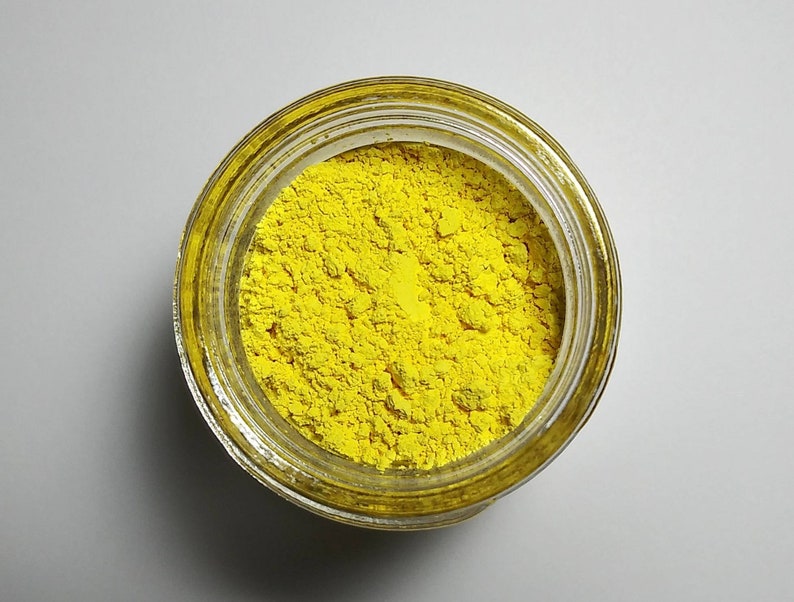 Puede incluir: Un tarro de vidrio transparente lleno de polvo amarillo brillante. El tarro est&aacute; abierto, revelando la sustancia finamente molida en su interior. El polvo tiene una apariencia texturizada y el tarro est&aacute; sobre una superficie blanca.