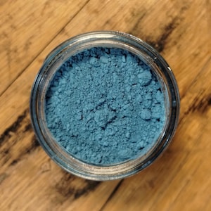 May include: A clear glass jar filled with a vibrant blue powder, set against a wooden surface. The powder has a fine, granular texture. The jar's rim is visible, and the wood grain adds a natural contrast to the blue pigment.