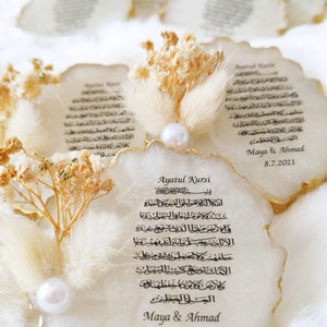 May include: White agate coaster with gold trim featuring the Arabic text of Ayatul Kursi, a verse from the Quran. The coaster is decorated with dried flowers and a pearl. The text "Maya & Ahmad 8.7.2021" is printed below the verse.