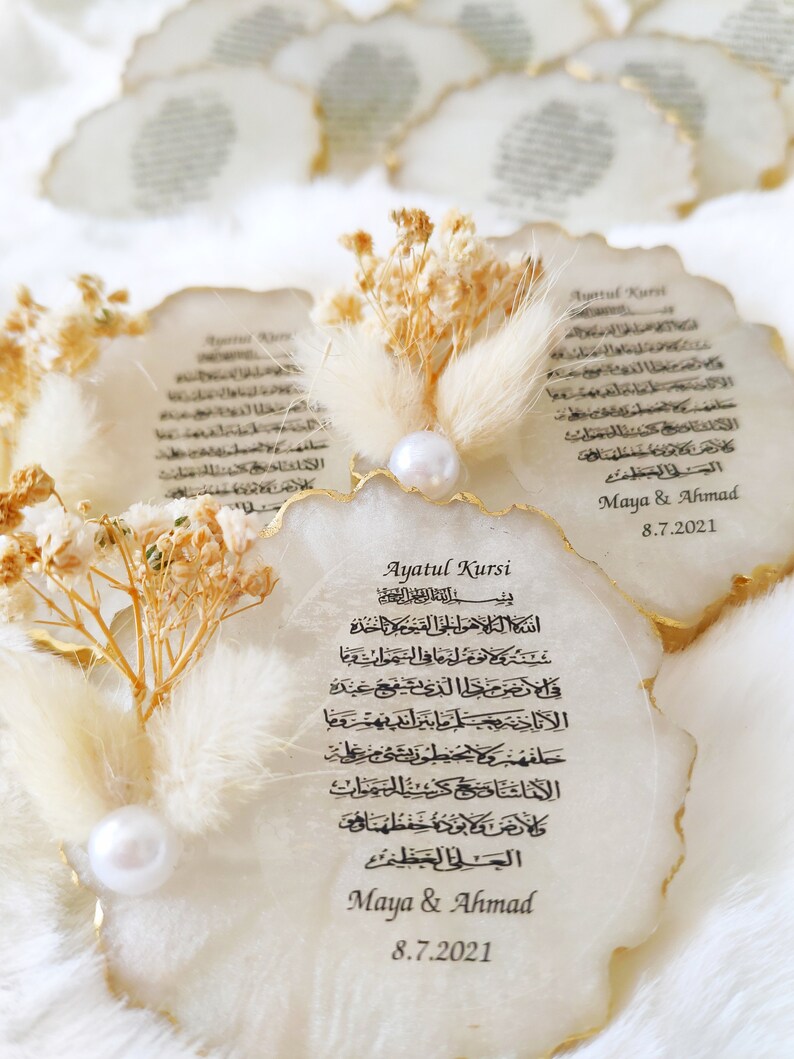May include: White agate coaster with gold trim featuring the Arabic text of Ayatul Kursi, a verse from the Quran. The coaster is decorated with dried flowers and a pearl. The text "Maya & Ahmad 8.7.2021" is printed below the verse.