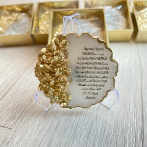 May include: A white, gold-flecked, resin plaque with a decorative, scalloped edge. The plaque features a traditional Islamic prayer, Ayatul Kursi, in Arabic script. The plaque is on a clear plastic stand.