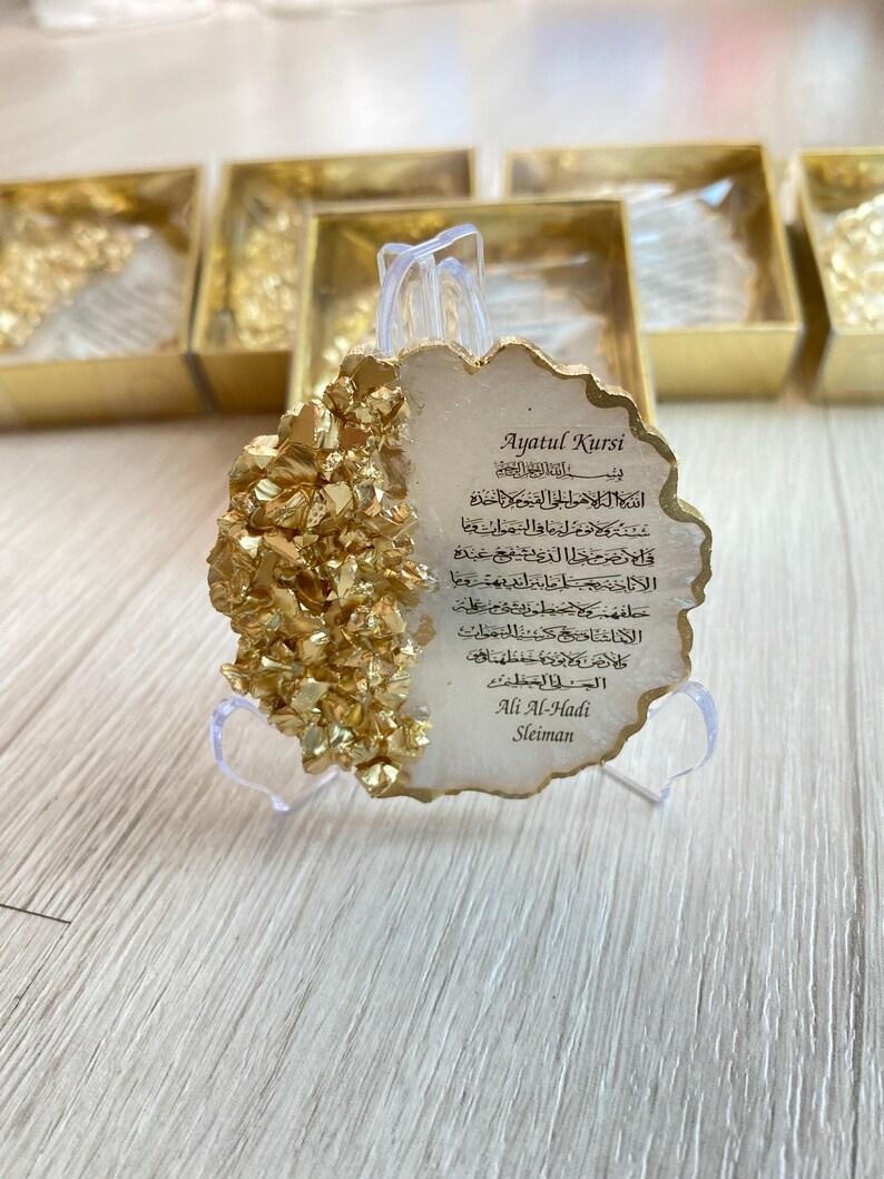 May include: A white, gold-flecked, resin plaque with a decorative, scalloped edge. The plaque features a traditional Islamic prayer, Ayatul Kursi, in Arabic script. The plaque is on a clear plastic stand.