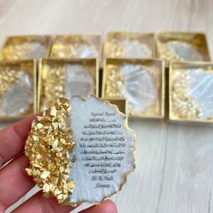 May include: A white and gold resin coaster with an Arabic inscription that reads "Ayatul Kursi" and includes the name "Ali Al-Hadi Sleiman".