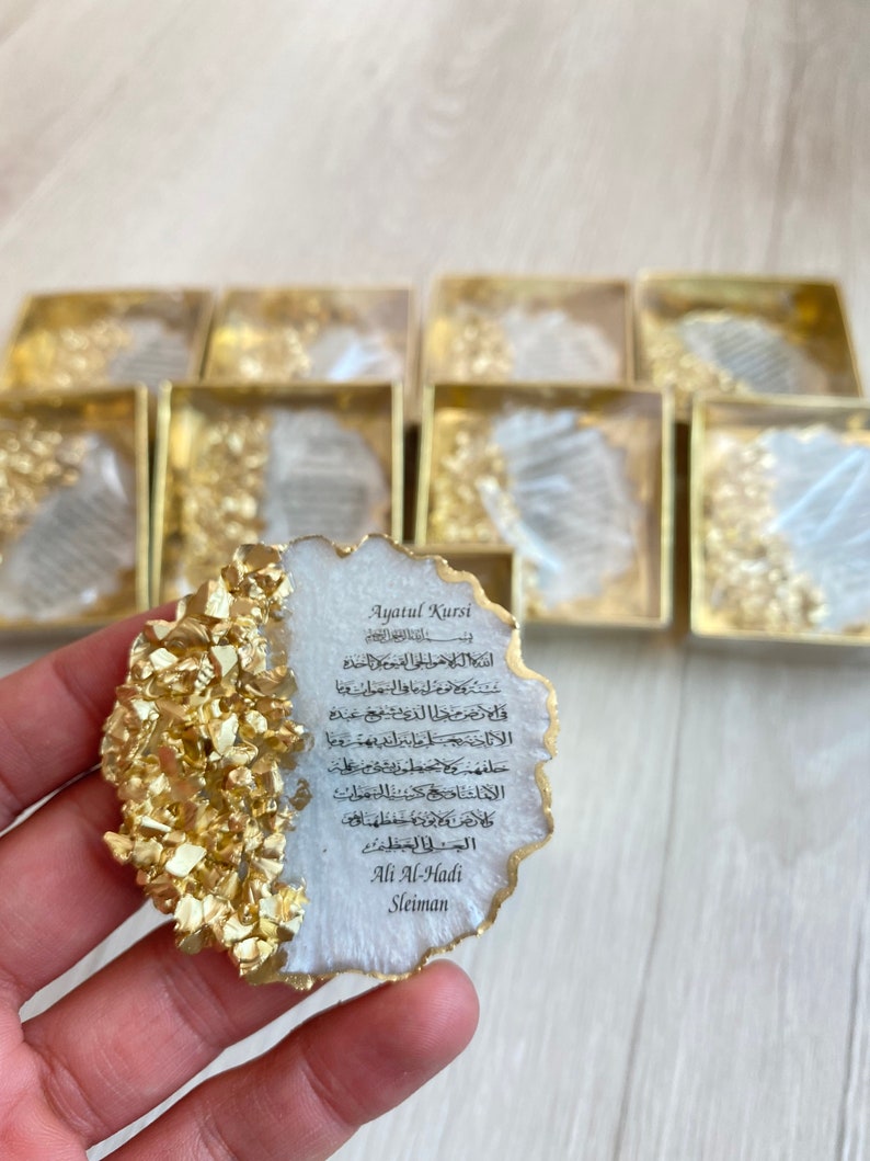 May include: A white and gold resin coaster with an Arabic inscription that reads "Ayatul Kursi" and includes the name "Ali Al-Hadi Sleiman".