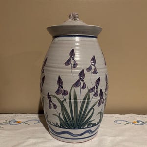 May include: A white ceramic jar with a lid, decorated with purple irises and green leaves. The jar has a blue and white stripe around the bottom.