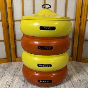 May include: A stackable, vintage kitchen canister set in yellow and orange. The set includes containers labeled "Tea", "Coffee", "Sugar", and "Flour". Each canister has a lid and a black label with white lettering.