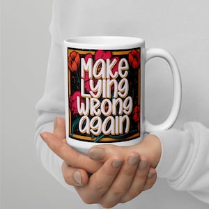 Może przedstawiać: Biały ceramiczny kubek z napisem "Make Lying Wrong Again" w stylizowanej czcionce. Projekt zawiera czerwone i pomarańczowe kwiatowe akcenty na czarnym tle, ze złotą obwódką.