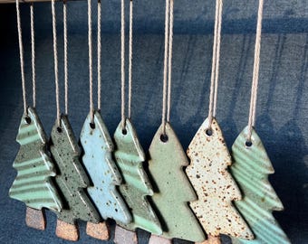 May include: Seven ceramic Christmas tree ornaments hanging from a wooden rod by twine strings. Each tree features a unique glaze, including shades of sage green, light blue, and speckled cream, with a distinct brown textured base on every piece.