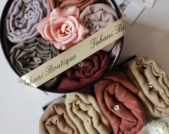 May include: A gift box filled with six rolled-up scarves in various shades of brown, beige, and gray. The box is decorated with a white ribbon that says "Tahani Boutique".