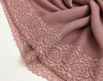 May include: A dusty rose-coloured fabric scarf featuring a wide, intricate lace border with a repeating floral rose pattern. The fabric has a soft, slightly textured appearance, and the lace trim follows the curved edge of the draped material.