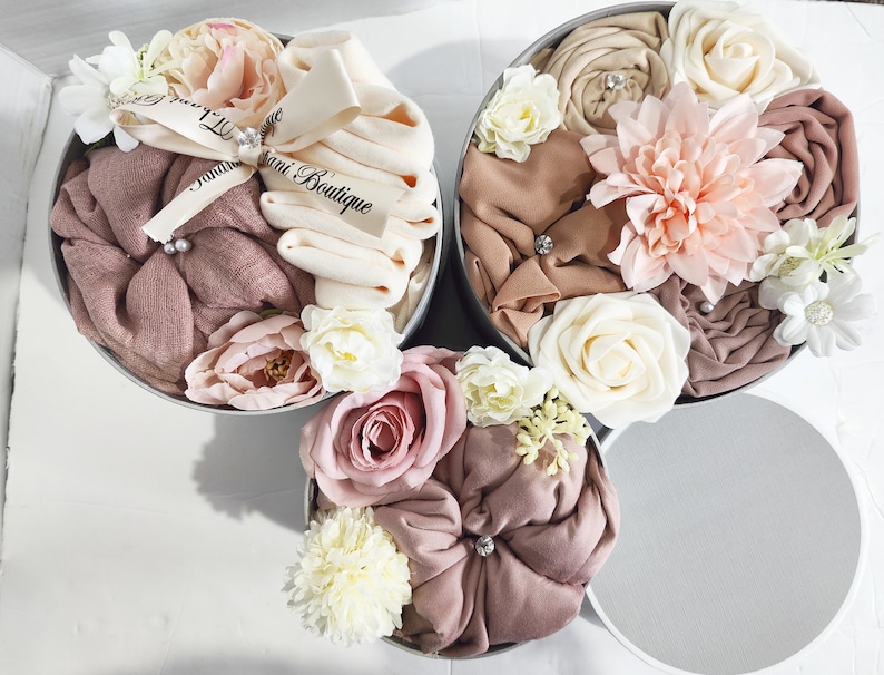 May include: Three round boxes filled with soft, pink and beige fabric, arranged in a pyramid shape. The boxes are decorated with delicate, artificial flowers in shades of pink, white, and cream. The top box has a white ribbon with the text "Jani Boutique" written on it.