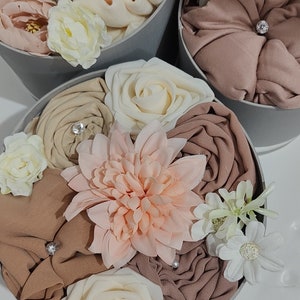 May include: A gift box filled with a variety of soft, brown, beige, and peach colored fabrics, decorated with white, peach, and cream colored artificial flowers. The box is gray and has a white label that says "Boutique".