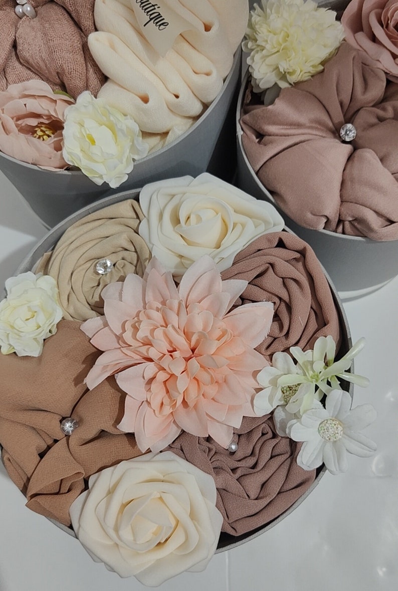May include: A gift box filled with a variety of soft, brown, beige, and peach colored fabrics, decorated with white, peach, and cream colored artificial flowers. The box is gray and has a white label that says "Boutique".
