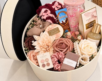 May include: A round cream-colored gift box filled with spa items including a pink tumbler with bow prints, Jonas & James body scrub and lotion, Options hot chocolate, maroon fuzzy socks, artificial flowers, a pearl necklace, and Tahani Boutique hair accessories.
