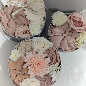 May include: Three round gift boxes filled with beige and brown fabric and artificial flowers. The boxes are decorated with white satin ribbon and the text "Sani Boutique".