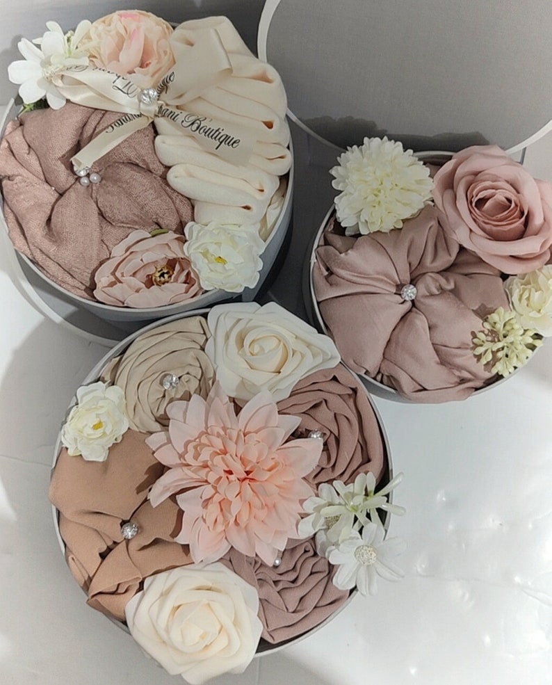 May include: Three round gift boxes filled with beige and brown fabric and artificial flowers. The boxes are decorated with white satin ribbon and the text "Sani Boutique".